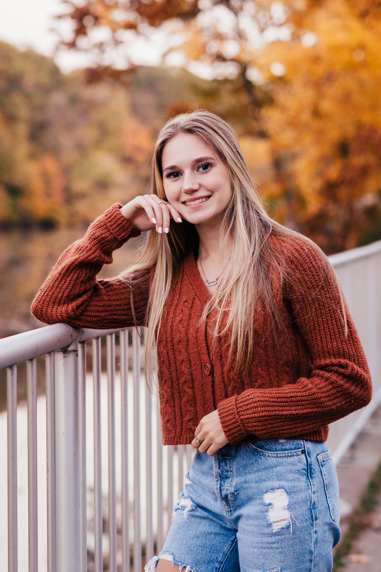Senior in a red sweater on a white railing with fall trees